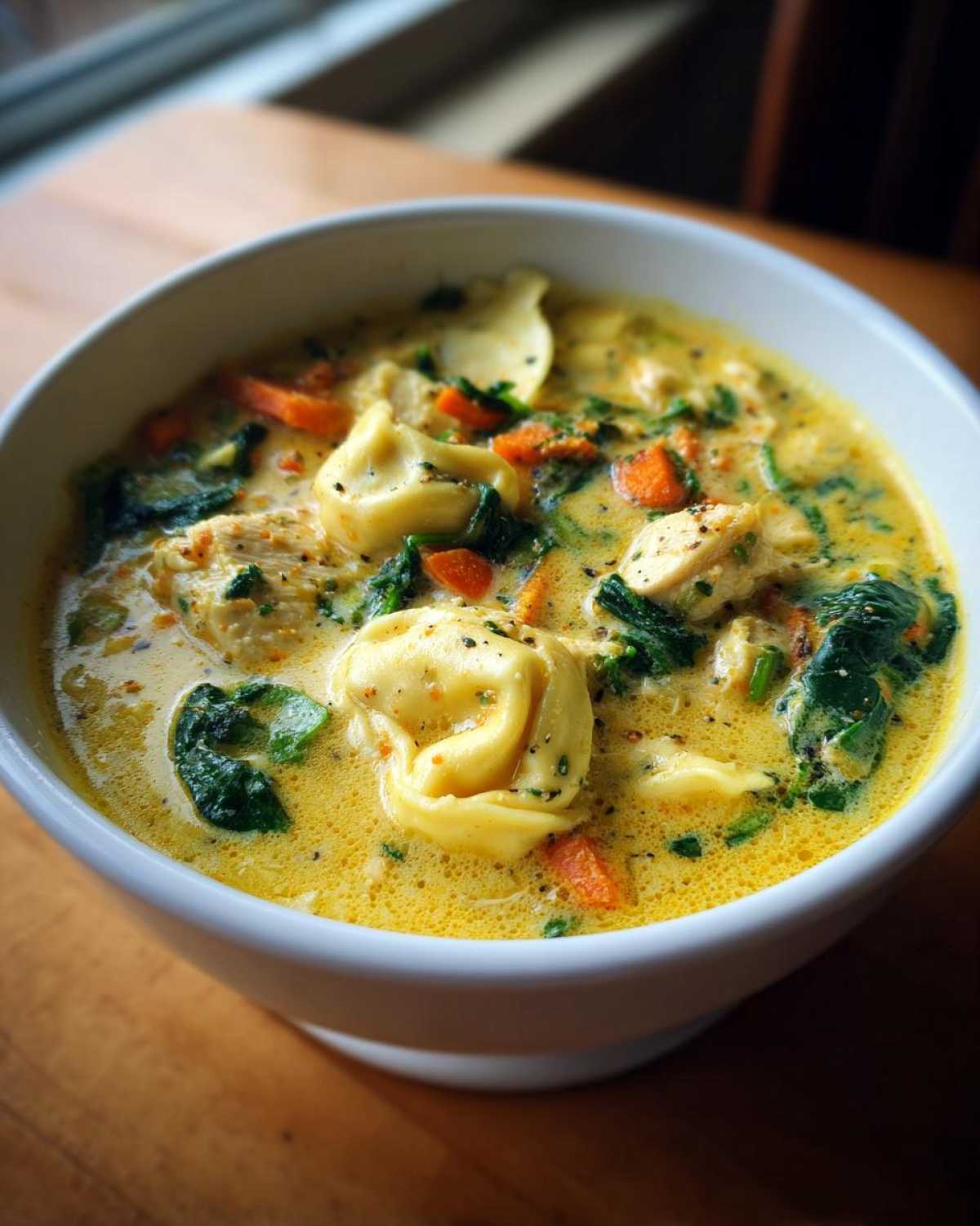 A close-up of a white bowl filled with rich, yellow creamy tortellini soup featuring pasta, chicken chunks, spinach, and carrots.