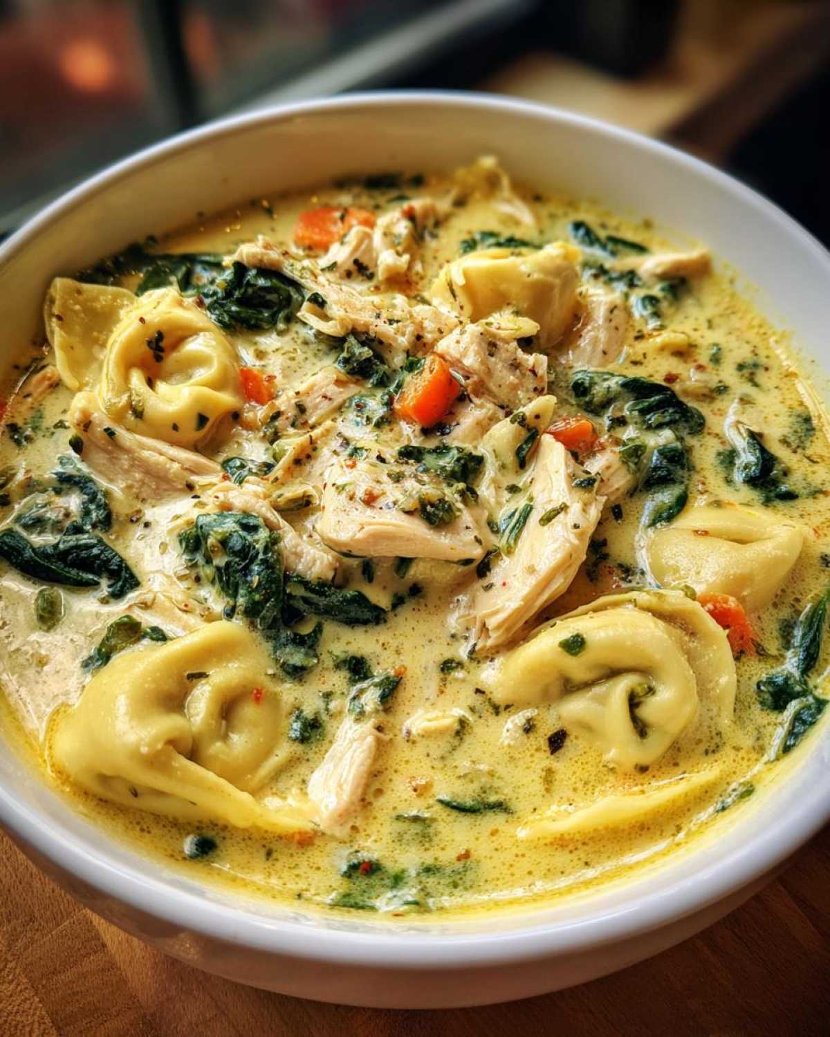 A close-up shot of a white bowl filled with rich, creamy tortellini soup featuring chunks of chicken, wilted spinach, and diced carrots.