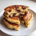 A stack of fluffy chocolate chip pancakes cut in half, showing melted chocolate chips inside.