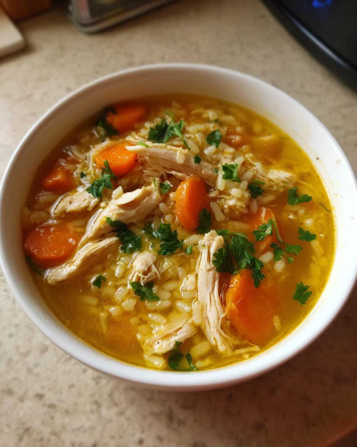 Close-up of a white bowl filled with Lemon Ginger Turmeric Chicken and Rice Soup, featuring shredded chicken, rice, carrots, and parsley.