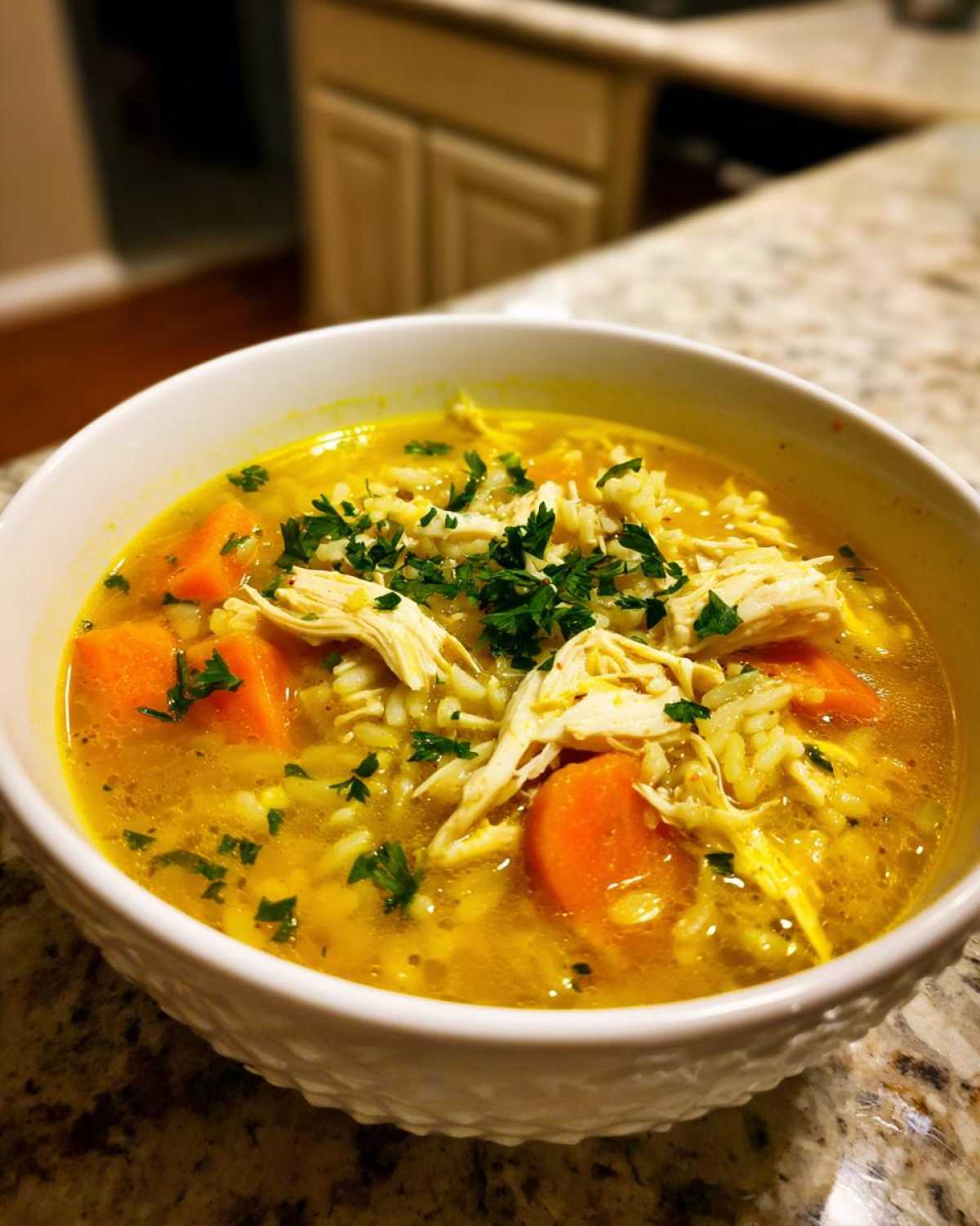 Close-up of a white bowl filled with vibrant yellow Lemon Ginger Turmeric Chicken and Rice Soup, topped with shredded chicken, carrots, and parsley.