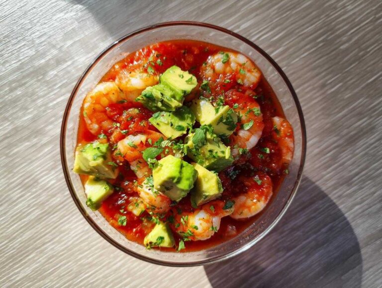 Overhead view of a glass bowl filled with Mexican shrimp cocktail, featuring shrimp in tomato sauce topped with diced avocado and cilantro.