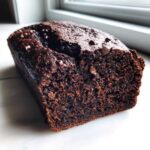 A close-up of a moist, dark chocolate bread loaf sprinkled with chocolate chips, resting on a white surface.