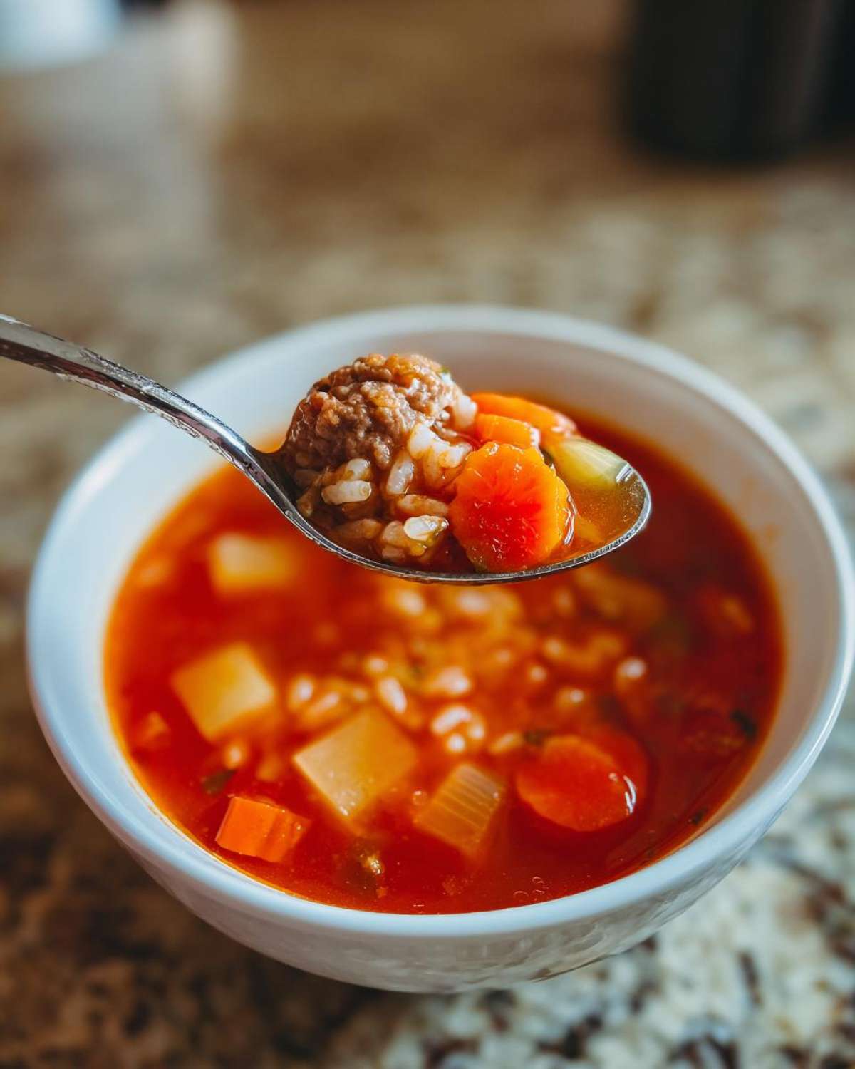 A spoonful of rich albondigas soup showing a meatball, rice, and sliced carrots being lifted from a white bowl.