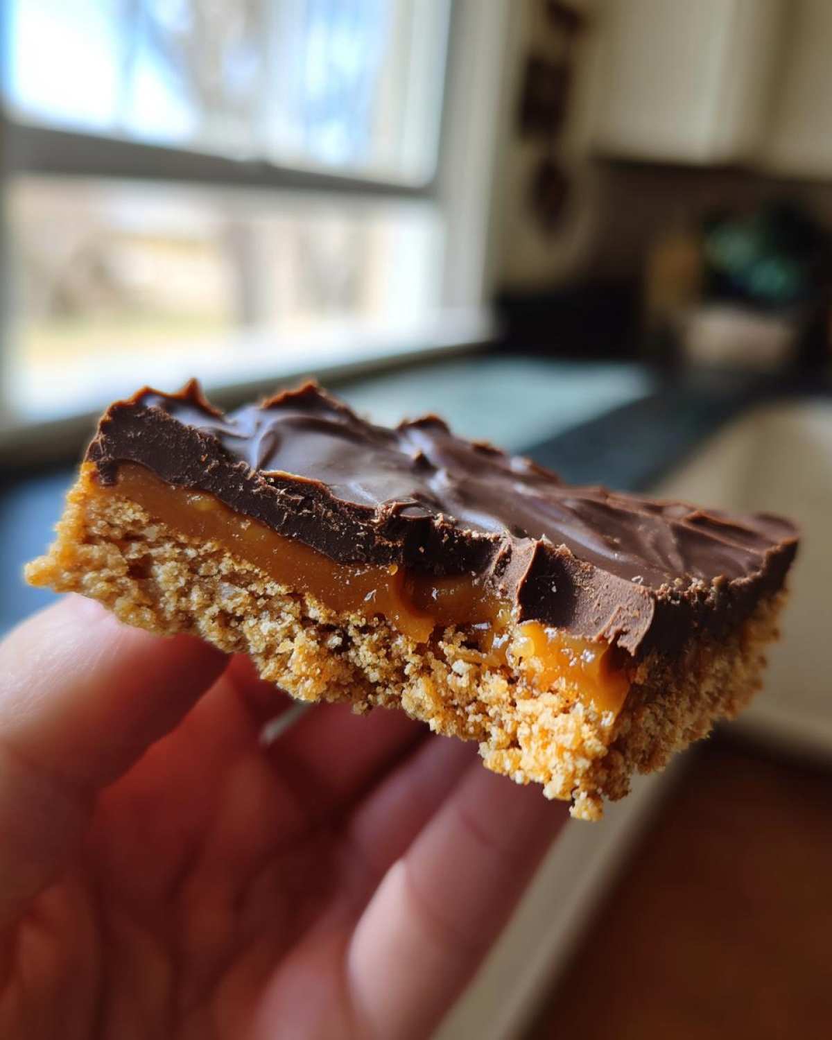 A hand holding a thick slice of graham cracker toffee with a crumbly base, gooey caramel layer, and melted chocolate top.