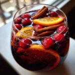 Close-up of a glass filled with dark red christmas sangria, garnished with fresh cranberries, orange slices, and cinnamon sticks.