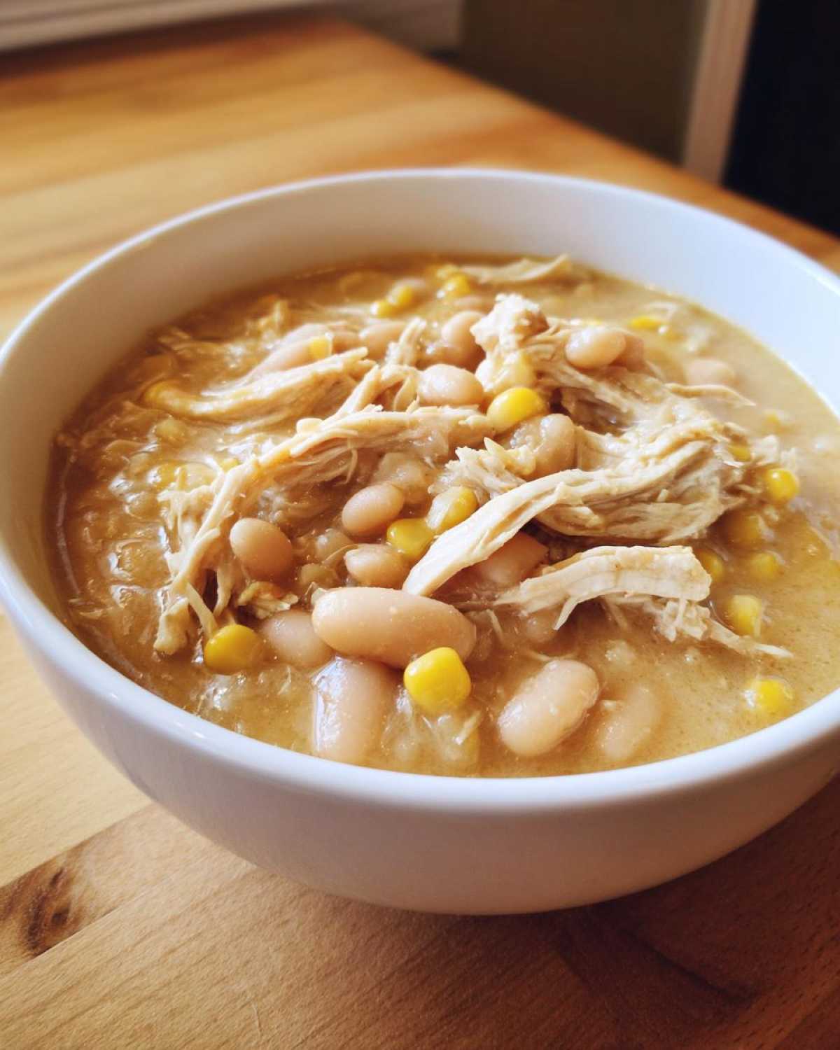 A close-up of a white bowl filled with creamy white chicken chili, featuring shredded white chicken, navy beans, and corn kernels.