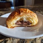 A golden brown chocolate croissant cut in half, revealing rich, melted chocolate filling inside, resting on a white plate.
