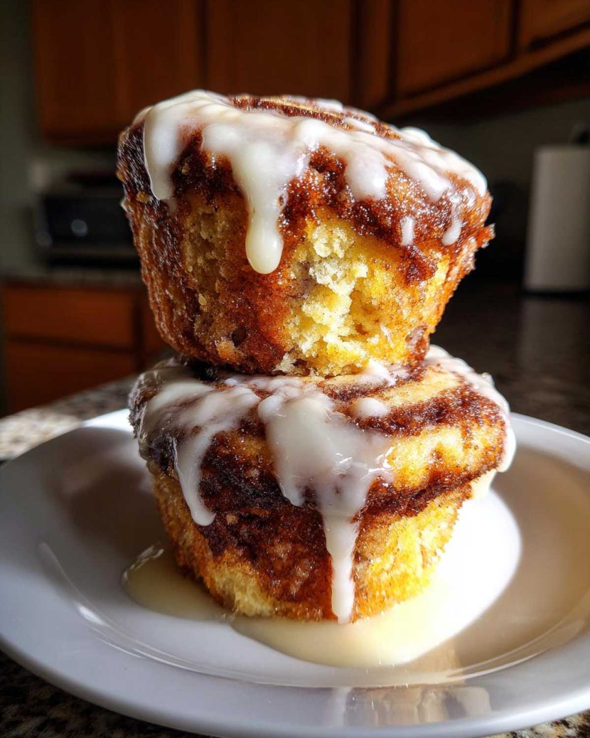 Two delicious cinnamon roll muffins stacked, dripping with sweet white icing, ready to eat.