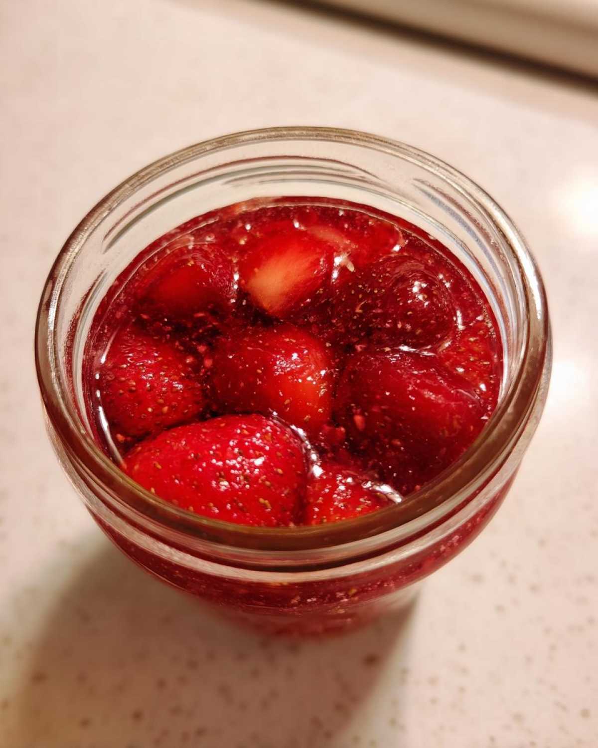 Overhead view of a glass jar filled with bright red strawberry freezer jam containing whole and halved strawberries.