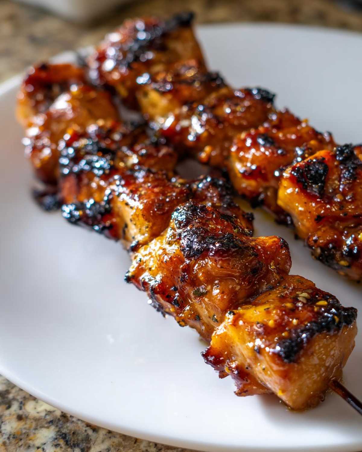 Two skewers of juicy, caramelized chicken skewers with charred edges resting on a white plate.