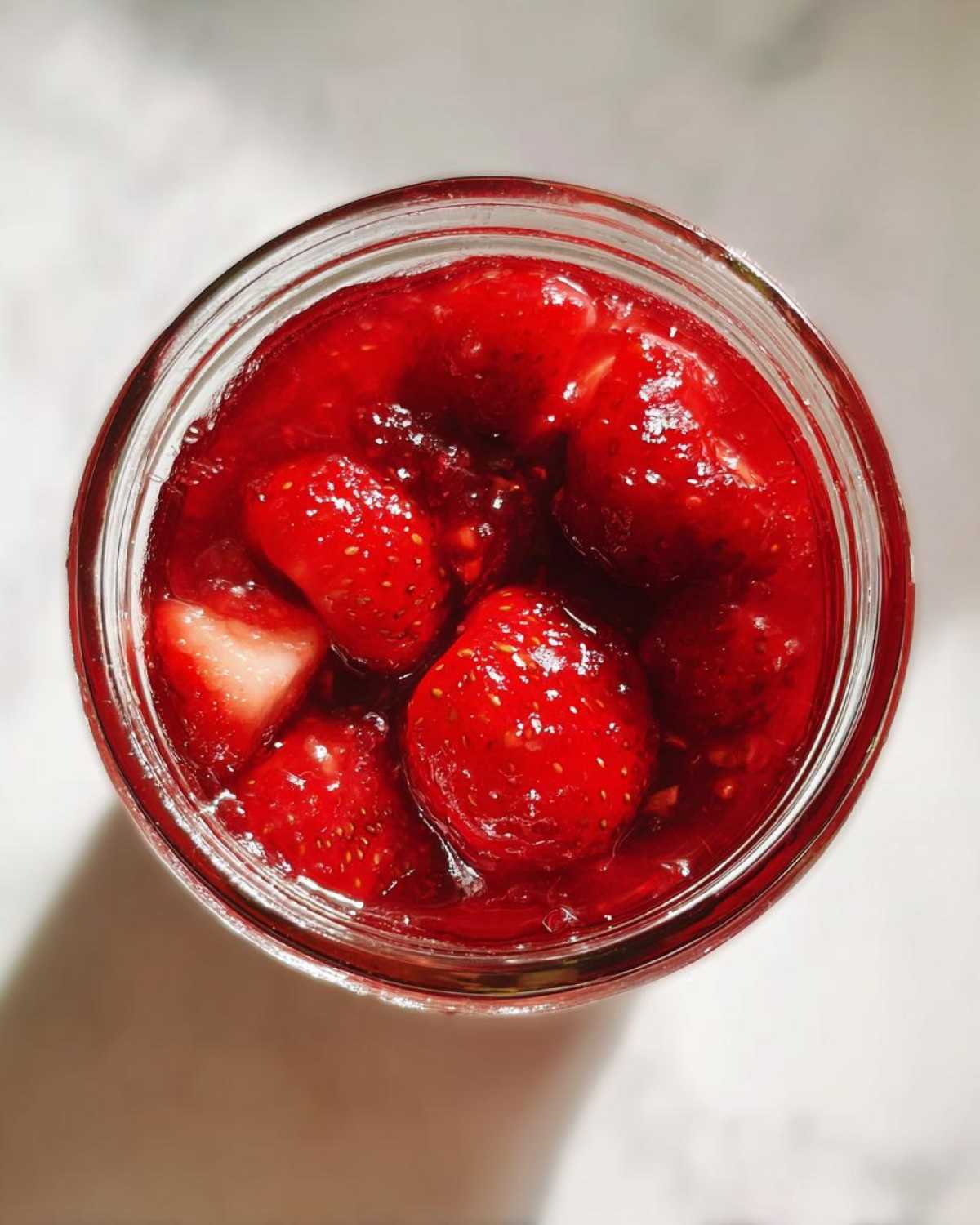 Overhead view of a glass jar filled with bright red strawberry freezer jam containing whole and halved strawberries.
