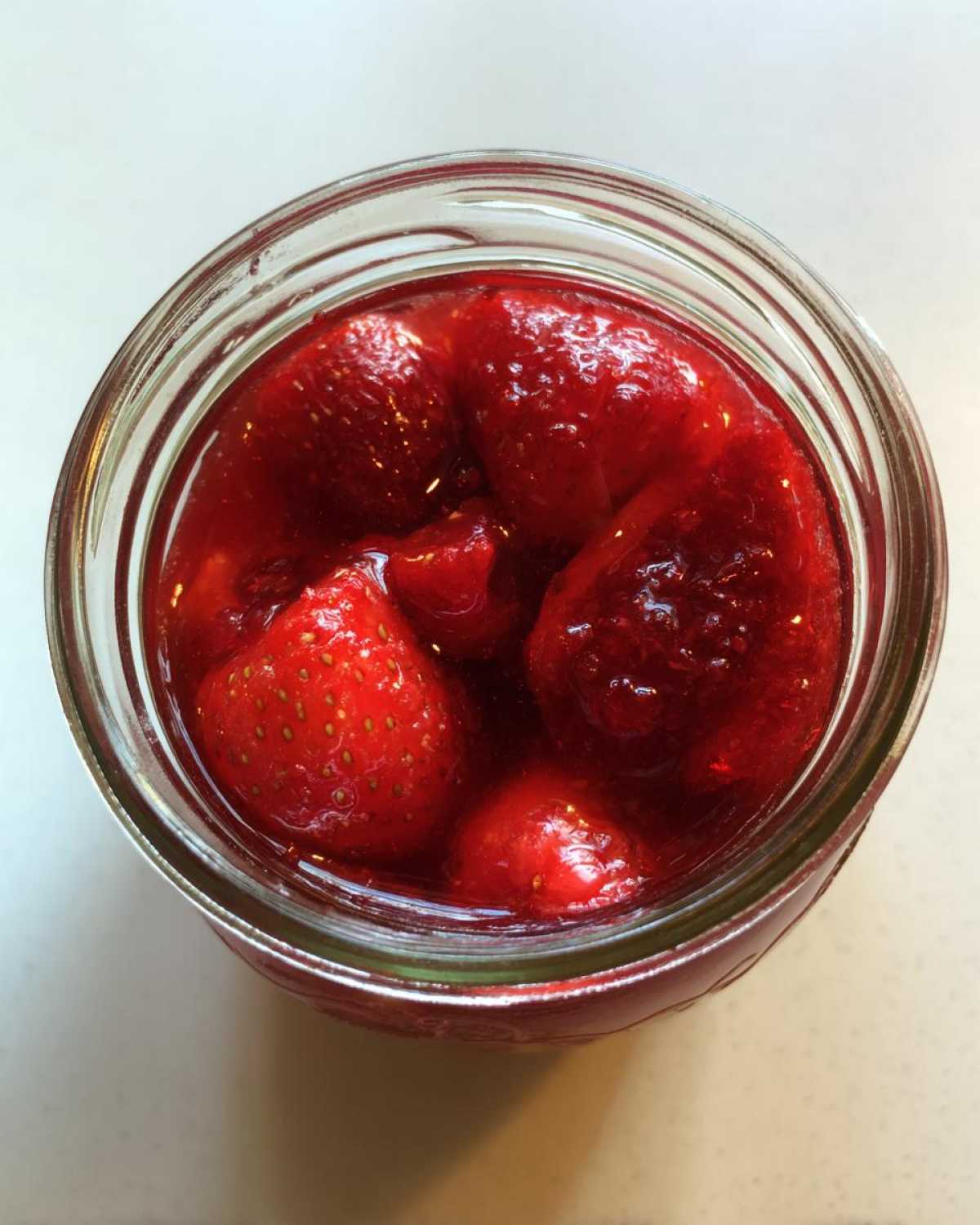 Overhead view of whole and halved strawberries suspended in bright red liquid inside a glass jar, ready for strawberry freezer jam.