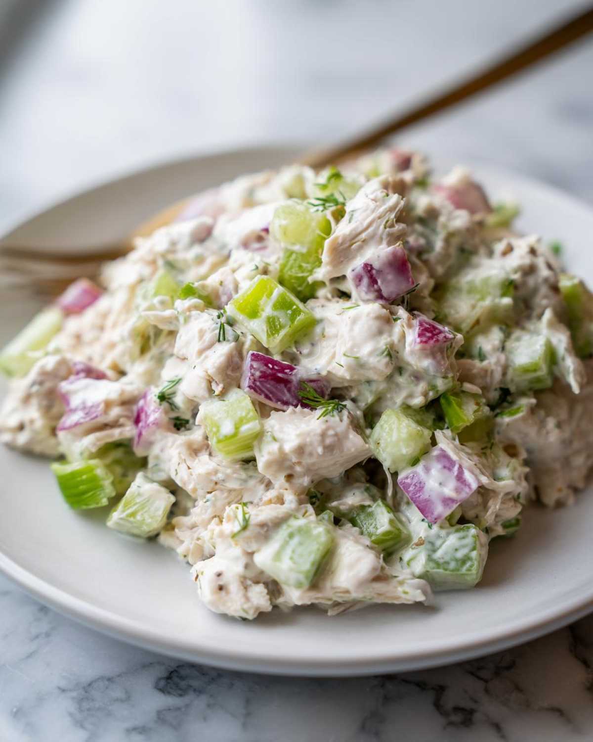 A close-up of creamy chicken protein salad mixed with diced celery and red onion on a white plate.