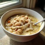 A close-up of a white bowl filled with creamy white chicken chili recipe, featuring shredded chicken and white beans.