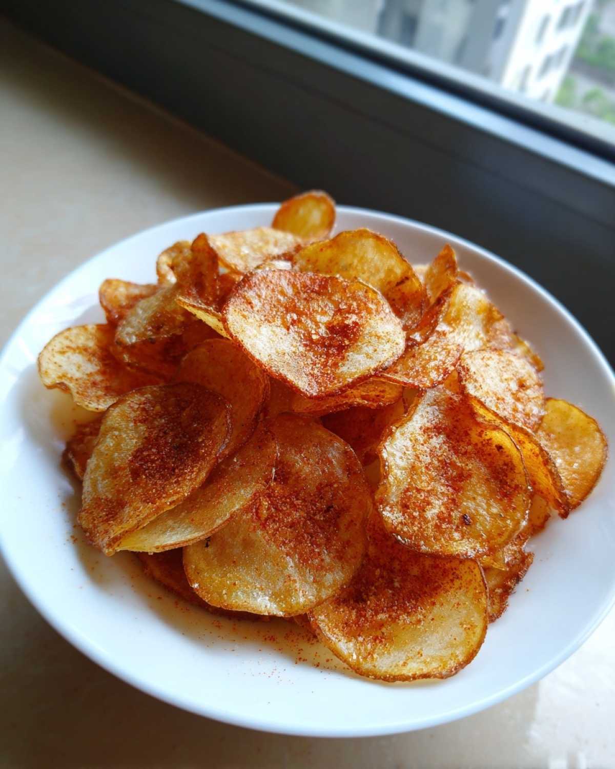 A white bowl filled with golden, crispy homemade potato chips dusted with red seasoning.