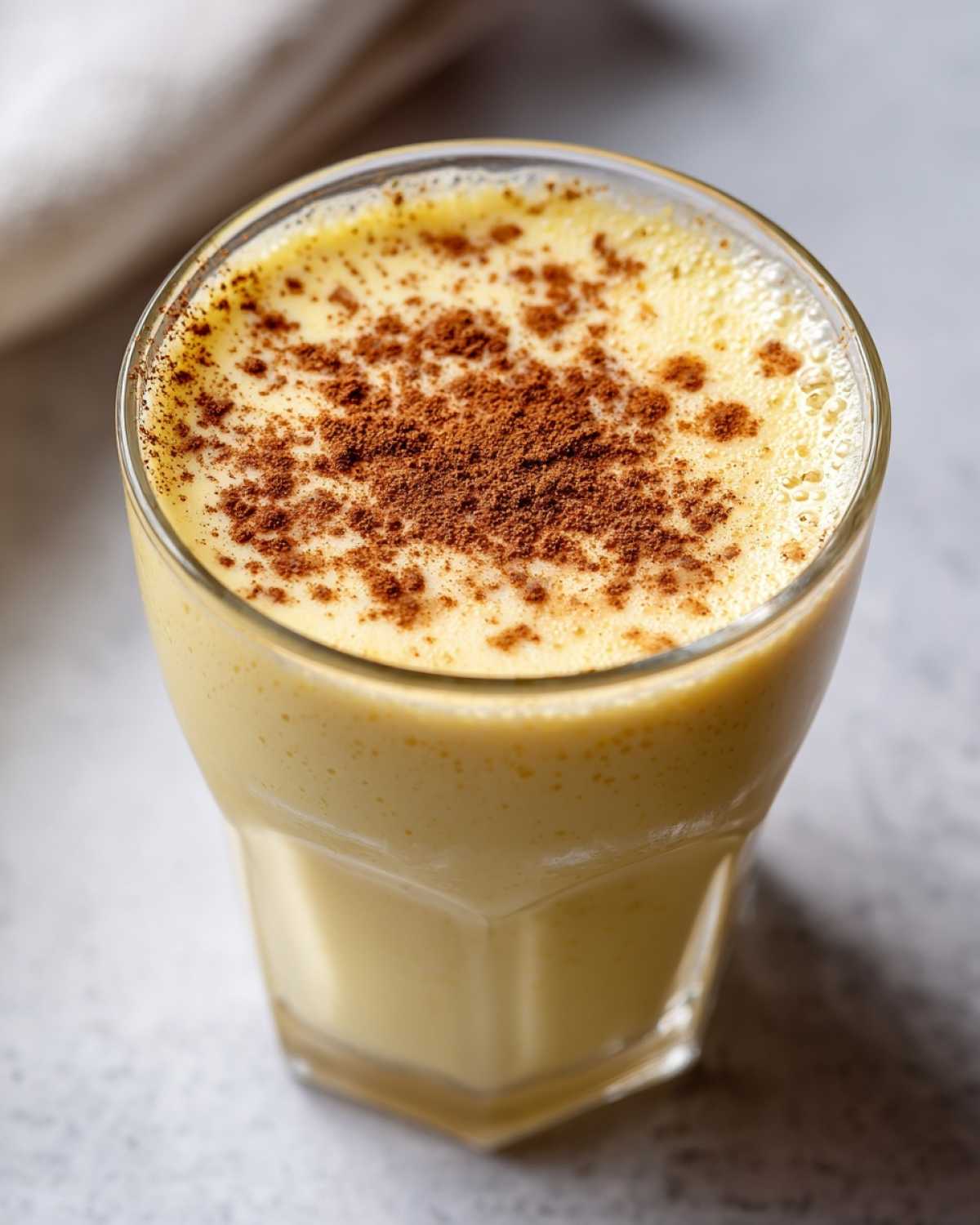 Close-up of a glass filled with creamy, yellow eggnog, topped with a dusting of cinnamon.