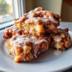 Close-up of a stack of golden brown, glazed apple fritter pieces piled on a white plate.