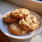 Four golden brown macadamia nut cookies stacked on a light ceramic plate, illuminated by sunlight.