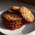 A stack of golden brown, crispy oatmeal lace cookies resting on a light-colored plate, illuminated by sunlight.