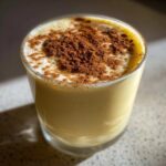 Close-up of a creamy, yellow glass of homemade eggnog topped with frothy foam and a heavy dusting of cinnamon.