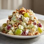 A mound of creamy Waldorf salad featuring green and red grapes, chopped celery, and topped generously with walnuts.