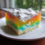 A square slice of rainbow jello poke cake showing layers of colorful gelatin, white cake, and topped with whipped cream.