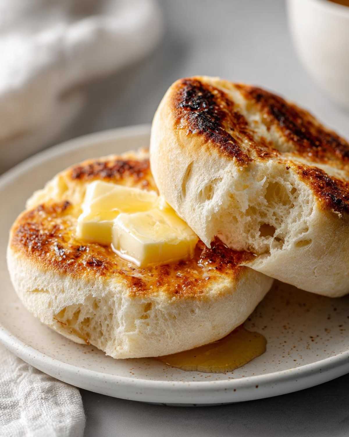 Close-up of toasted english muffins split open, topped with melting butter and honey on a speckled plate.