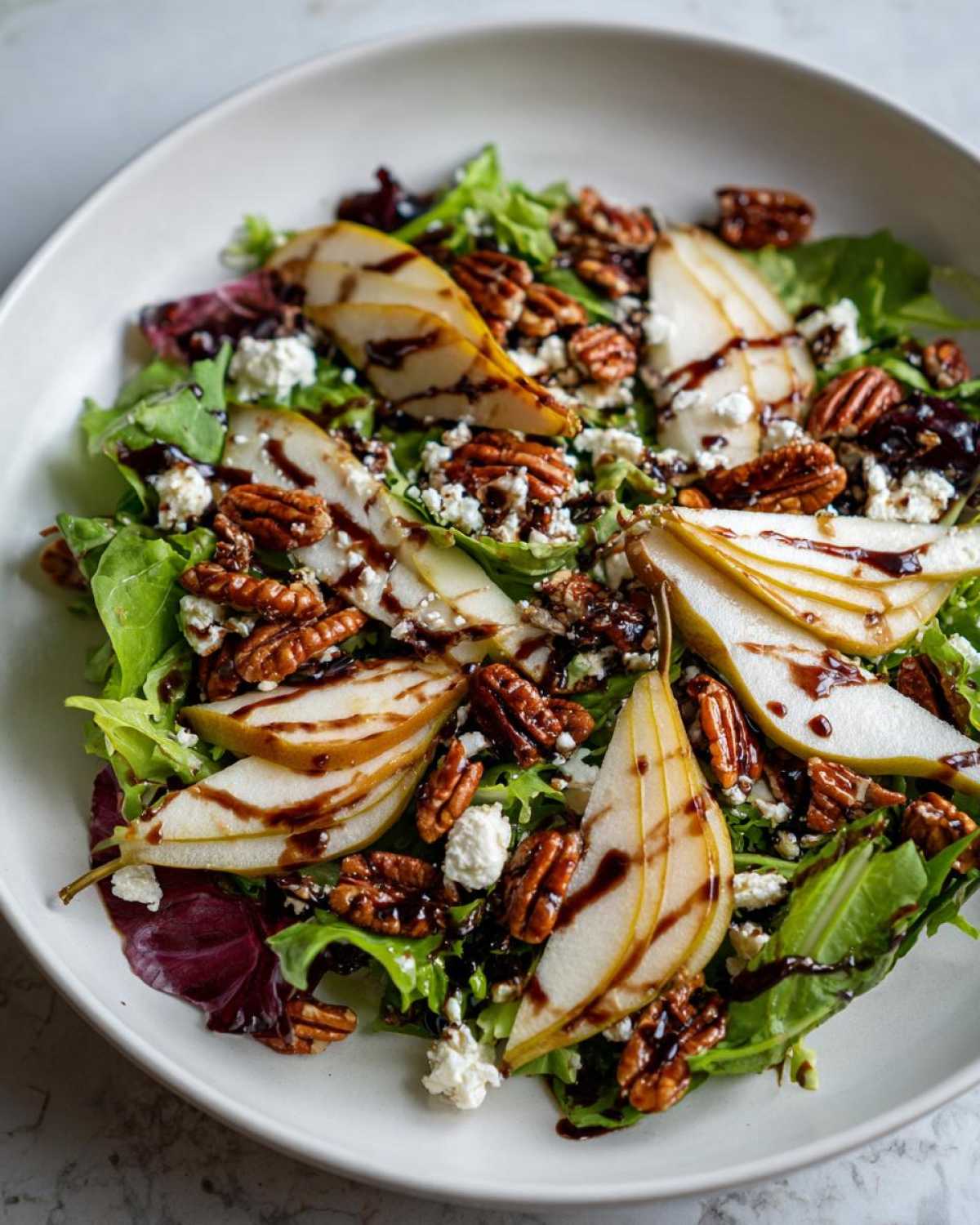A vibrant pear salad featuring sliced pears, candied pecans, crumbled cheese, and a balsamic glaze over mixed greens.