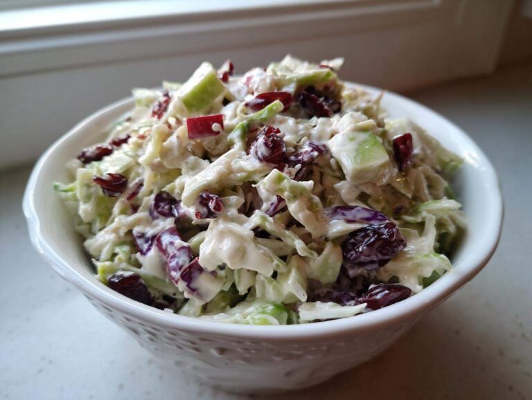 A close-up of creamy apple cranberry coleslaw mixed with shredded cabbage and dried cranberries, served in a white bowl.