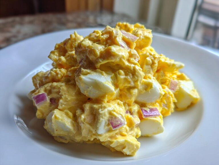 A mound of rich, yellow creamy egg salad featuring visible chunks of hard-boiled egg white and small pieces of red onion, served on a white plate.