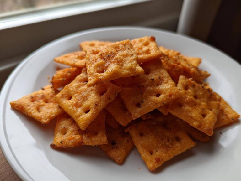A pile of freshly baked homemade healthy cheez-its, seasoned with pepper, resting on a white plate near a window.