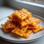 A close-up stack of golden, square homemade healthy cheez-its seasoned with paprika on a light gray plate.