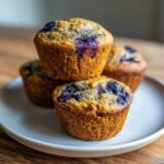A stack of four golden brown Banana blueberry protein muffins topped with baked blueberries on a light ceramic plate.