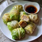 A plate of steamed cabbage dumplings with spicy soy dip, showing the savory meat filling inside a cut dumpling.