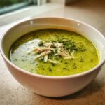 A bright green bowl of chilled zucchini soup with chicken shreds and fresh herbs on top.