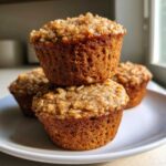 A stack of three golden brown High fiber apple cinnamon oatmeal muffins topped with oats, resting on a white plate.