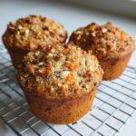 Close-up of three golden-brown High fiber morning glory muffins cooling on a wire rack, topped with nuts and shredded carrots.