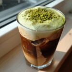 A tall glass of iced Pistachio tiramisu latte with layers of coffee, milk, green cold foam, and crushed pistachios on top.